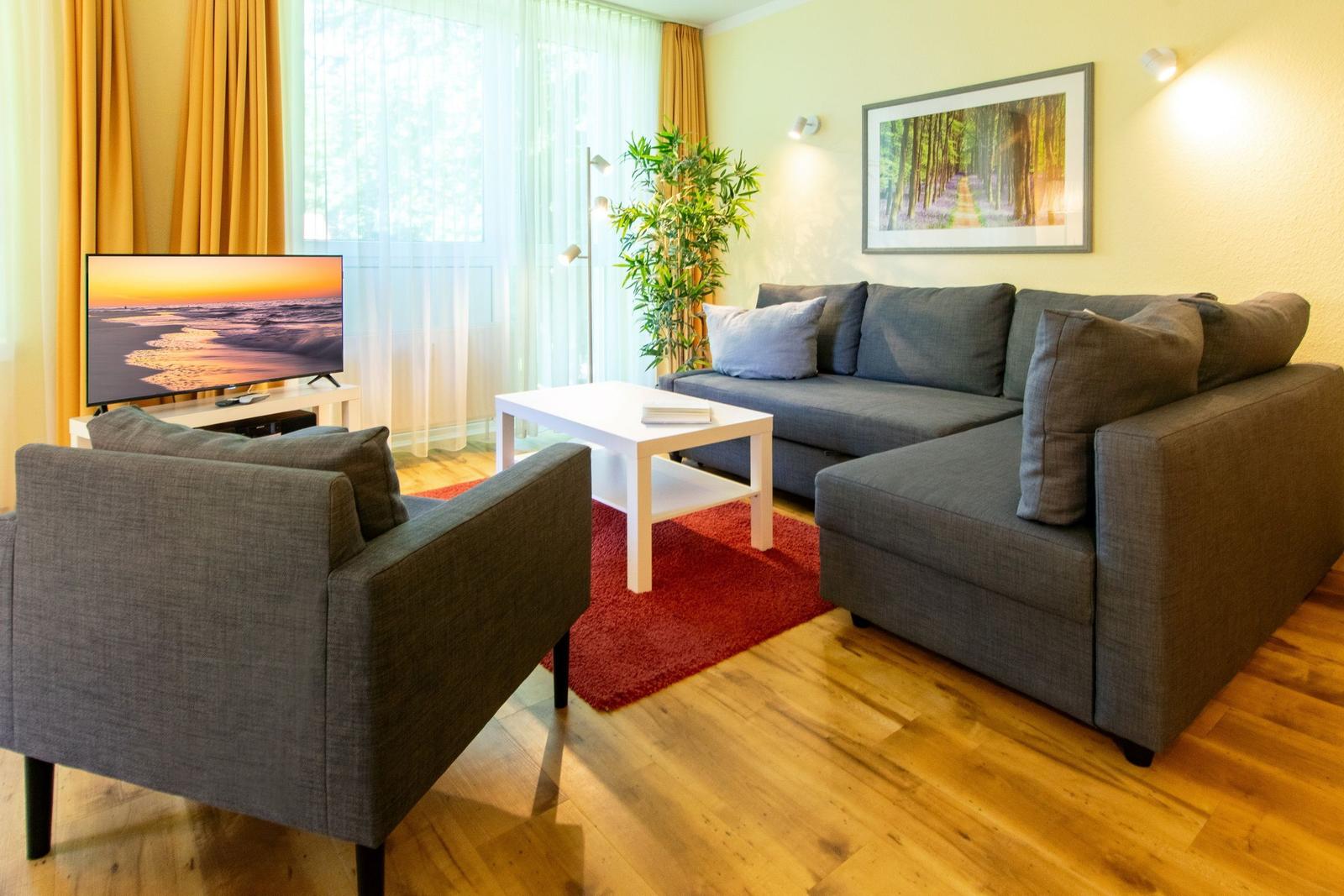 Wohnzimmer mit Sofa, Sessel, Couchtisch und Fernseher. Holzboden und Fenster mit Vorhängen.