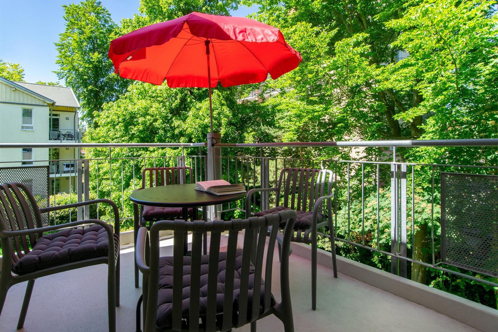 Terrasse mit Tisch, vier Stühlen und rotem Sonnenschirm. Umgeben von grünen Bäumen.