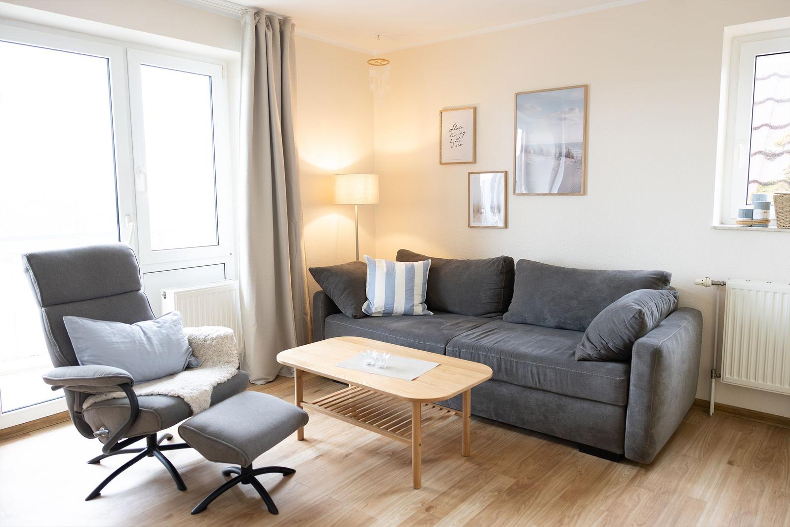 Wohnzimmer mit grauem Sofa, Sessel und Holztisch. An der Wand hängen Bilder.