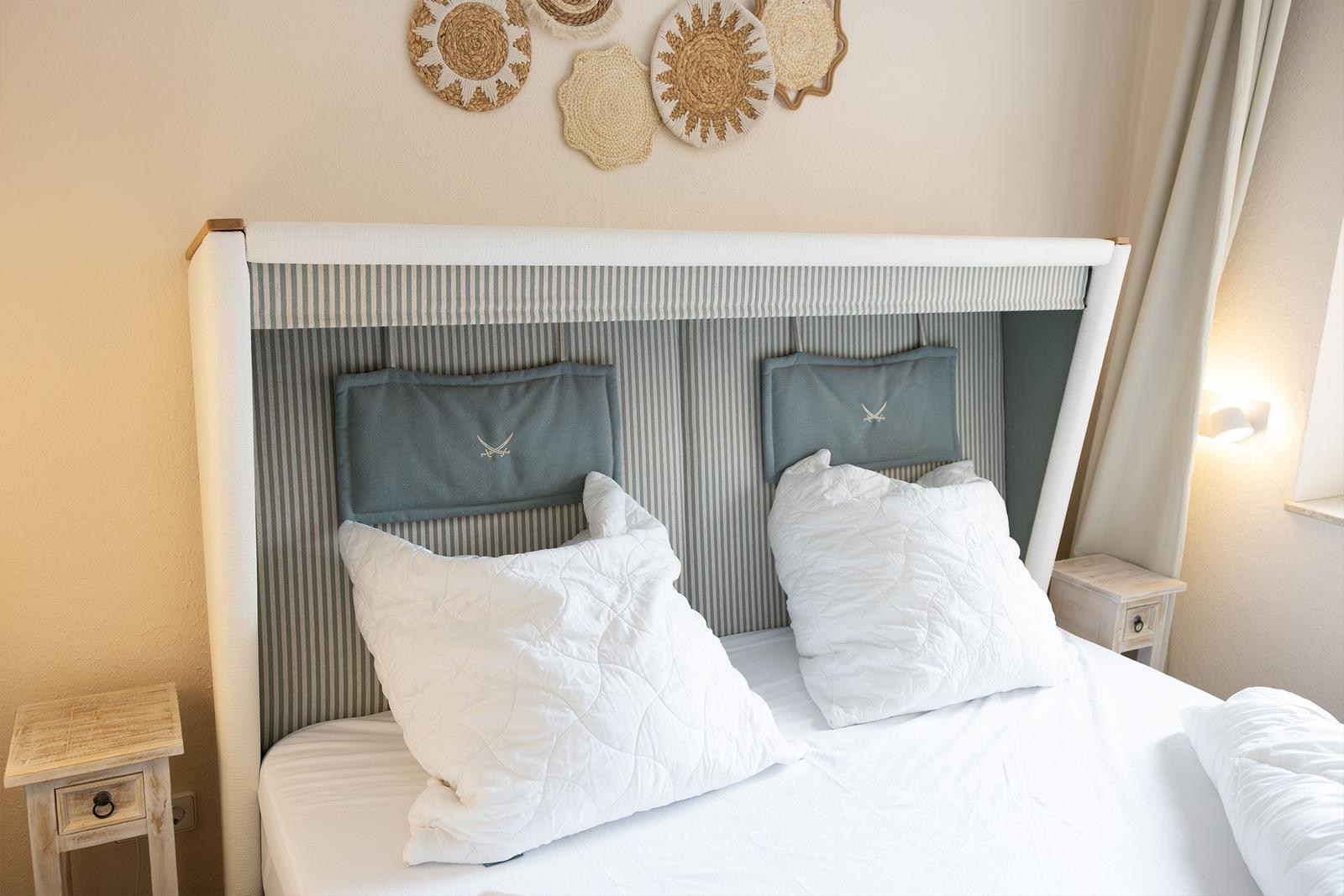 Ein Bett mit weißer Bettwäsche und zwei Kissen. An der Wand hängen raffinierte Wanddekorationen.