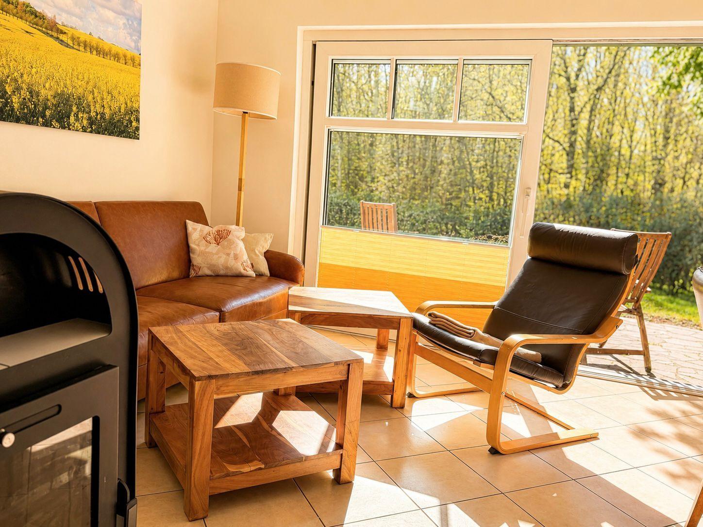 Living area with leather sofa, wooden table, and chair. Entrance to terrace with view into the forest.