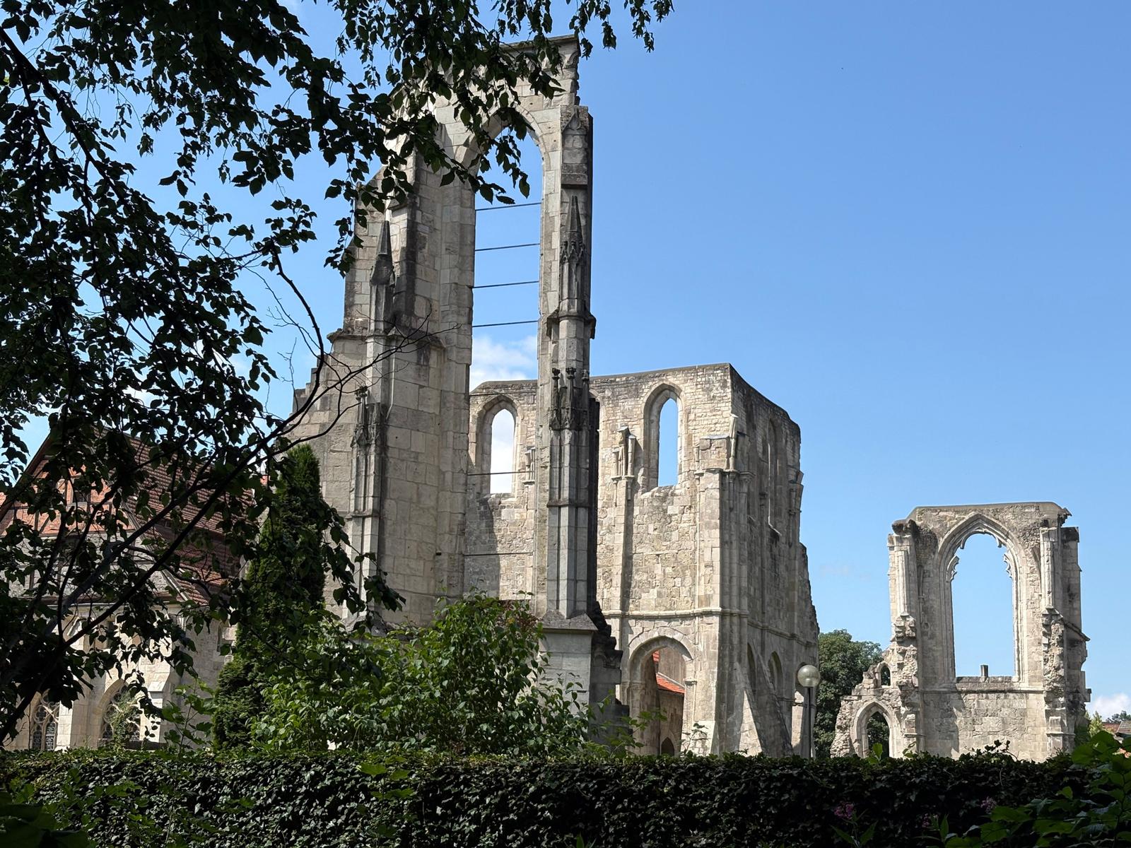 Ruinen einer gotischen Kirche unter blauem Himmel, umgeben von Bäumen und Sträuchern.