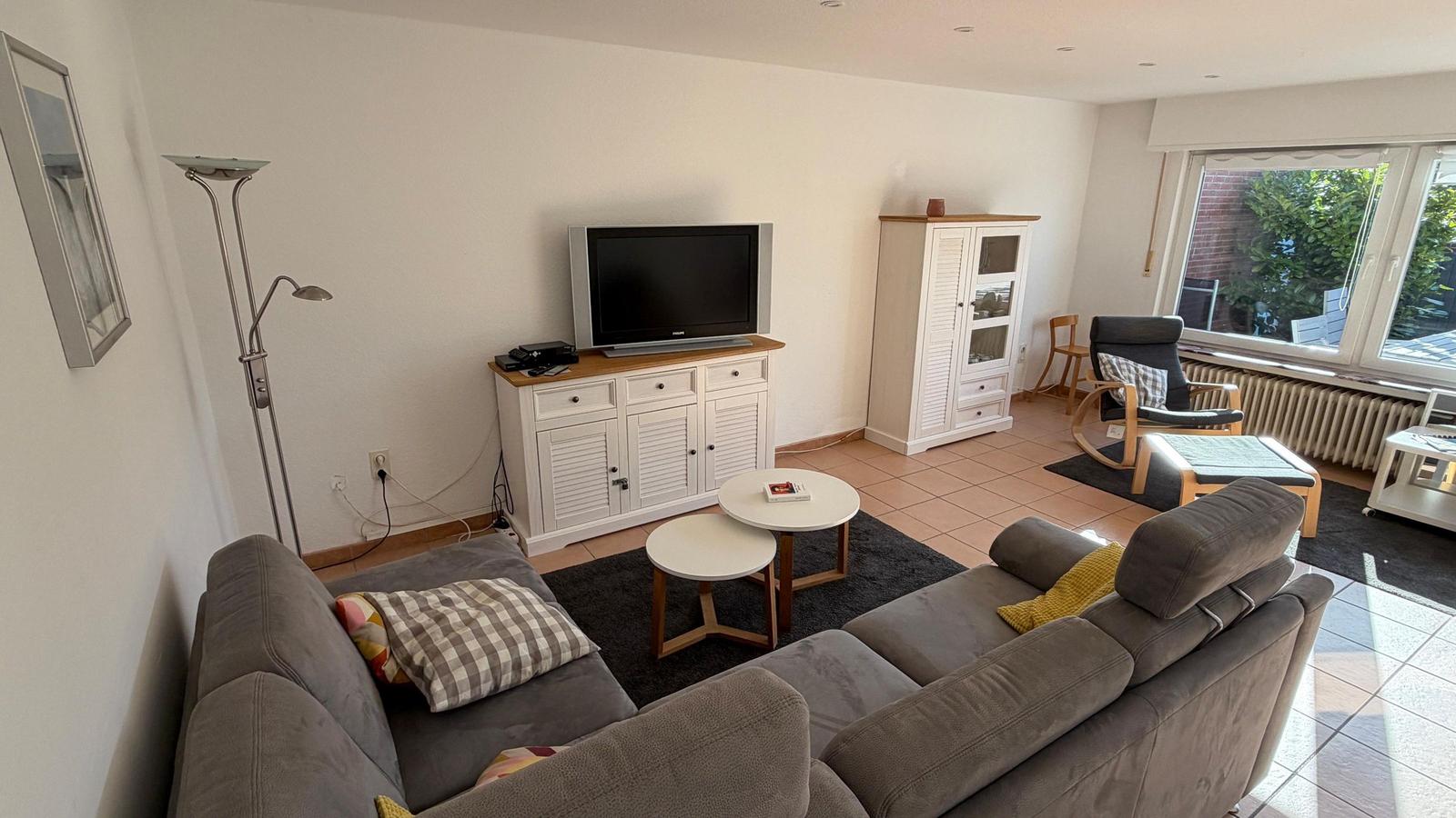 Wohnzimmer mit Sofa, Fernseher und Sitzgelegenheiten. Fensterblick auf Garten.