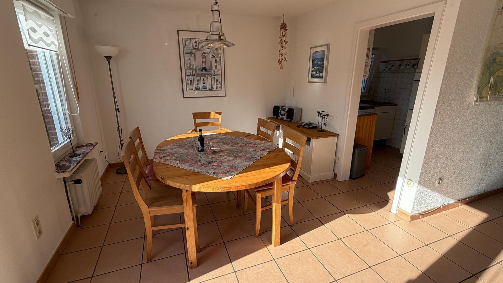 Esszimmer mit runden Tisch, vier Stühlen und Fenster. Küche ist angrenzend.