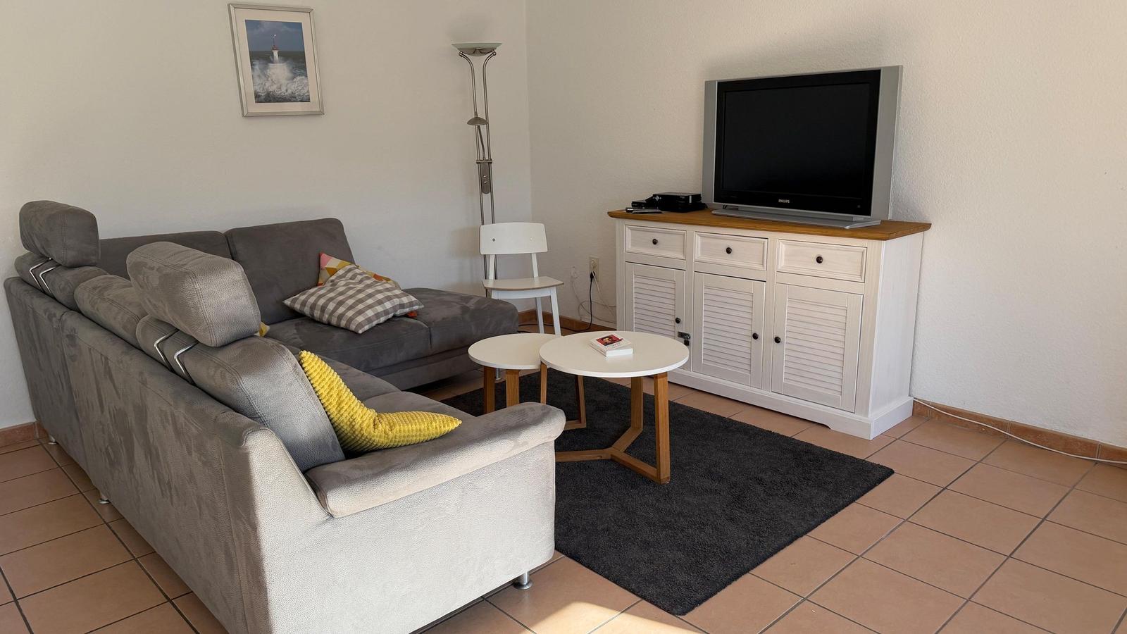 Ein Wohnzimmer mit grauem Sofa, zwei runden Tischen und einem Fernseher auf einem weißen Schrank.