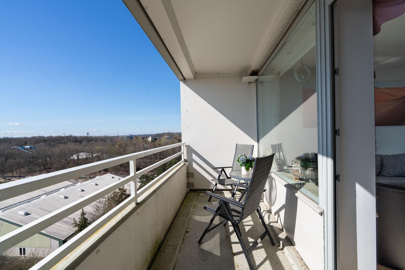 Balcony with two chairs and a small table. View over rooftops and trees under clear sky.