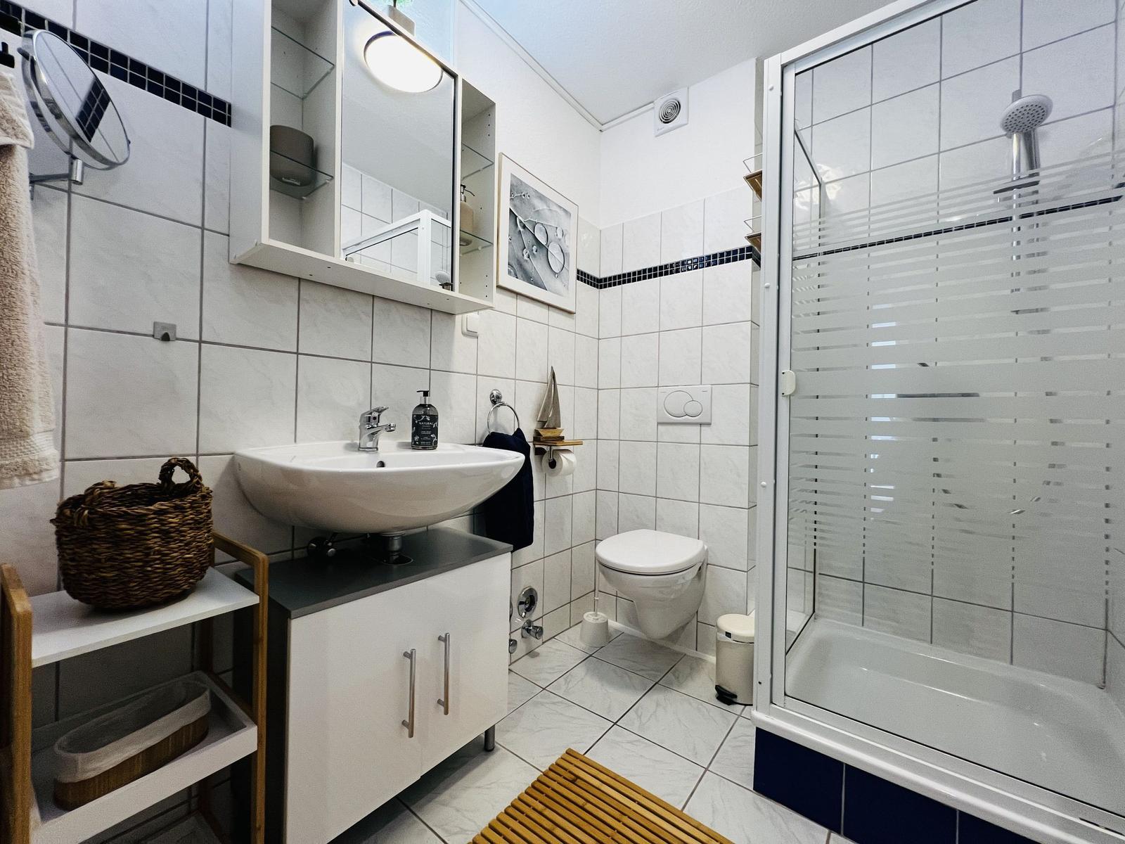 Badezimmer mit Dusche, Waschbecken und Toilette. Weiße Fliesen und Holzmatte.