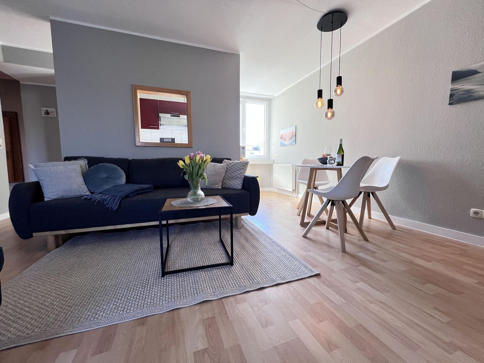 Wohn- und Essbereich mit Sofa, Tisch und Stühlen. Holzboden und Wandspiegel sichtbar.