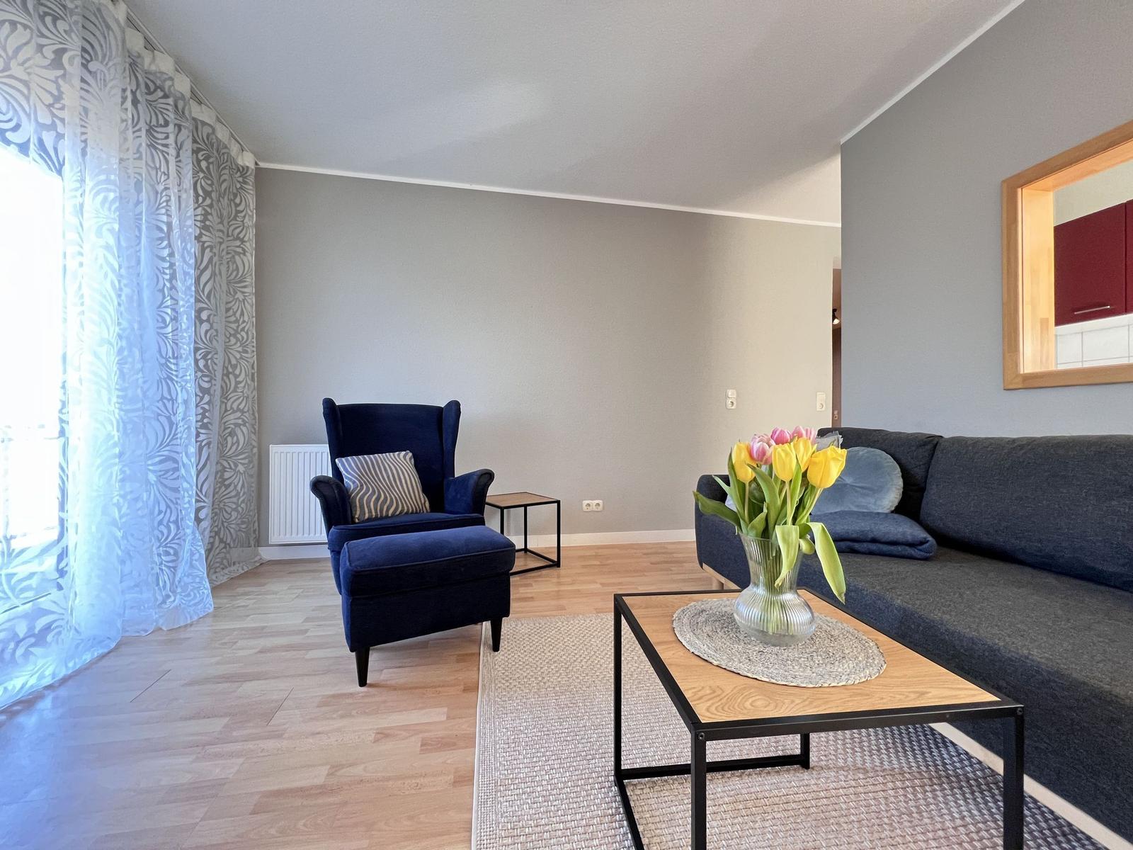 Wohnzimmer mit Sofa, Sessel, Tisch und Blumen.