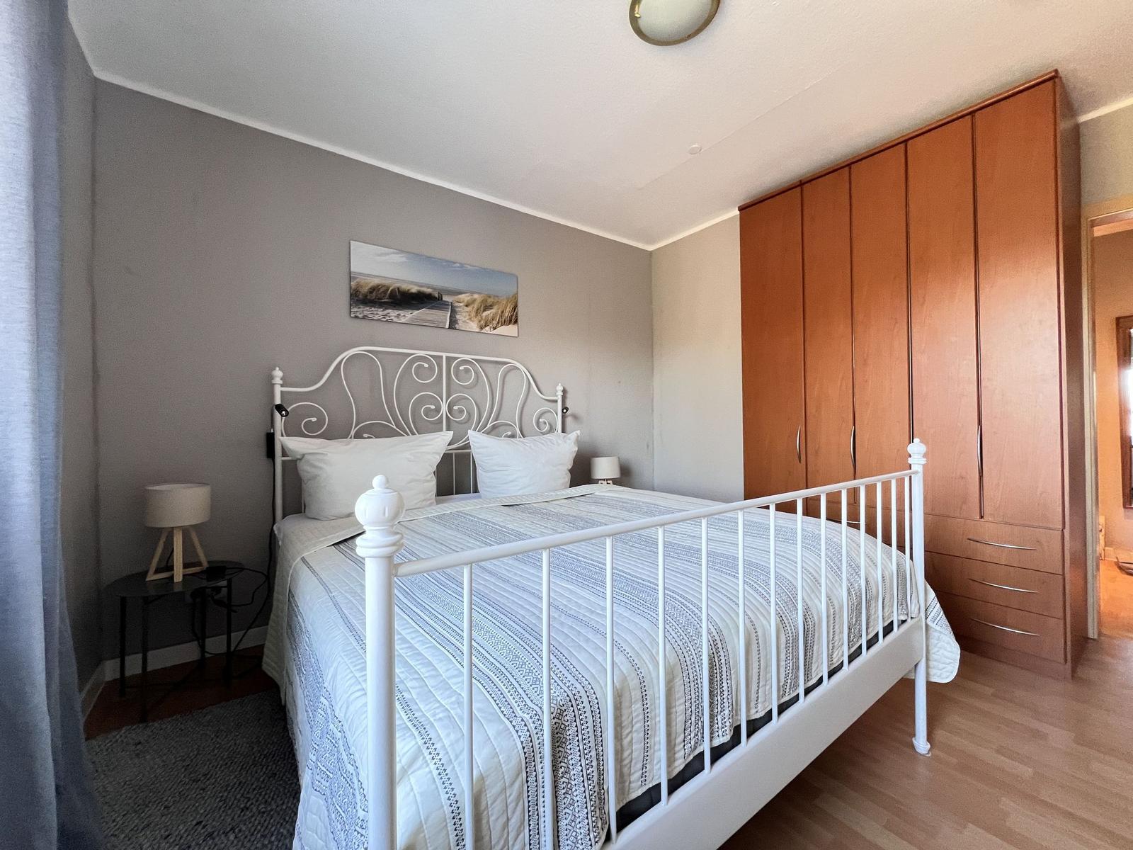 Schlafzimmer mit weißem Metallbett, Holzschrank und Wandbild.