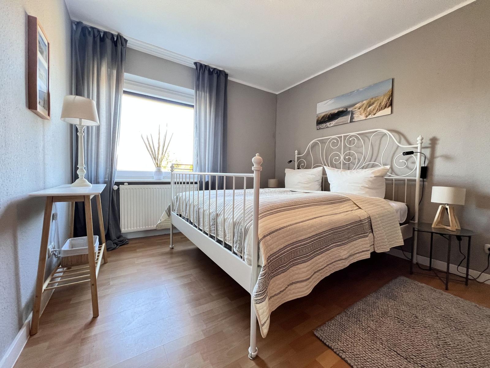 Schlafzimmer mit weißem Metallbett, Holzboden und Fenster mit Vorhängen.