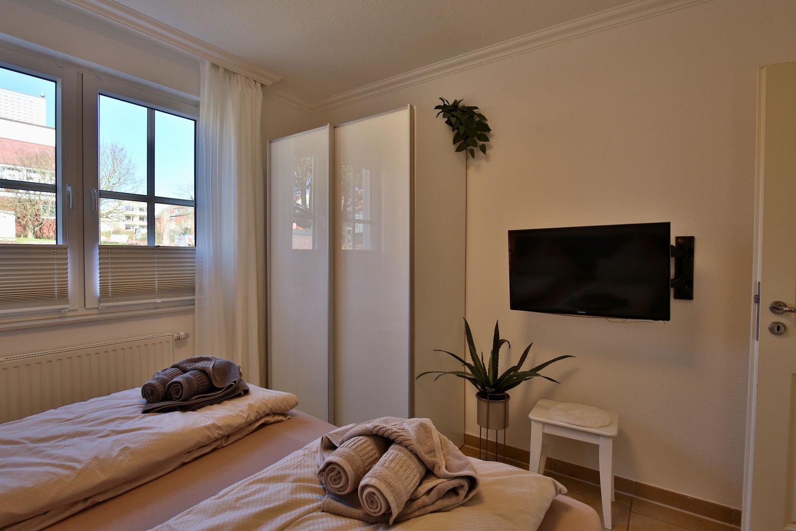 Two beds with towels, window, wardrobe, and wall-mounted TV.