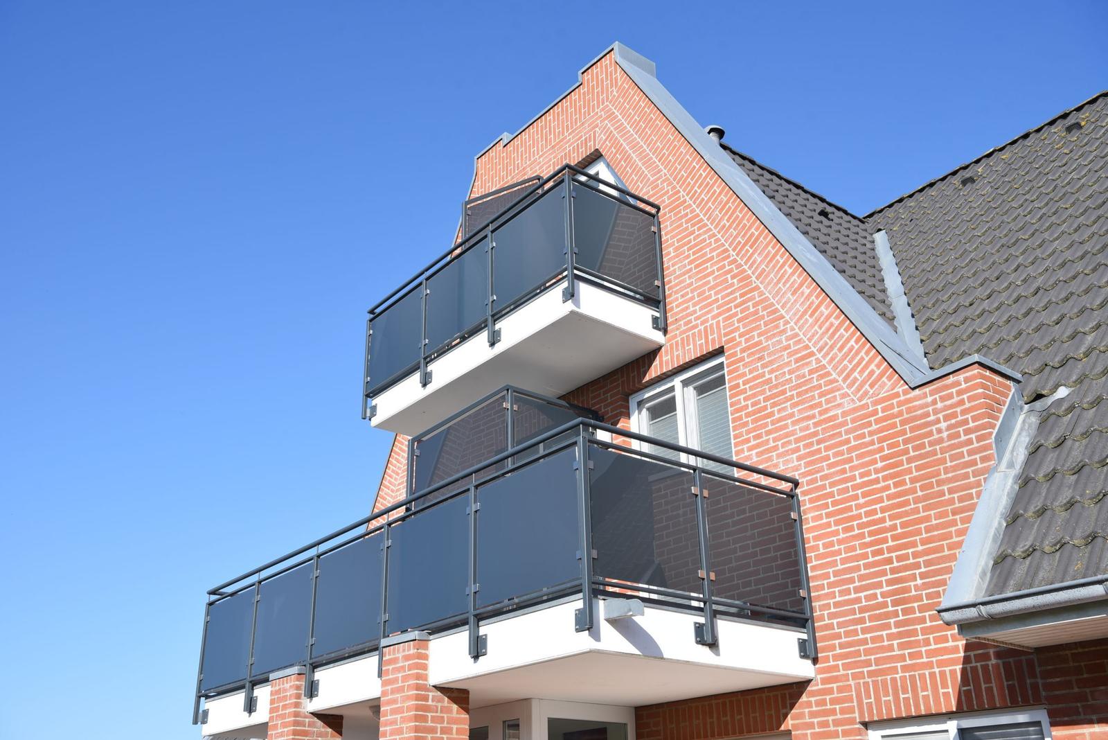 Zwei Balkone mit Glasgeländer an einem Backsteinhaus unter blauem Himmel.