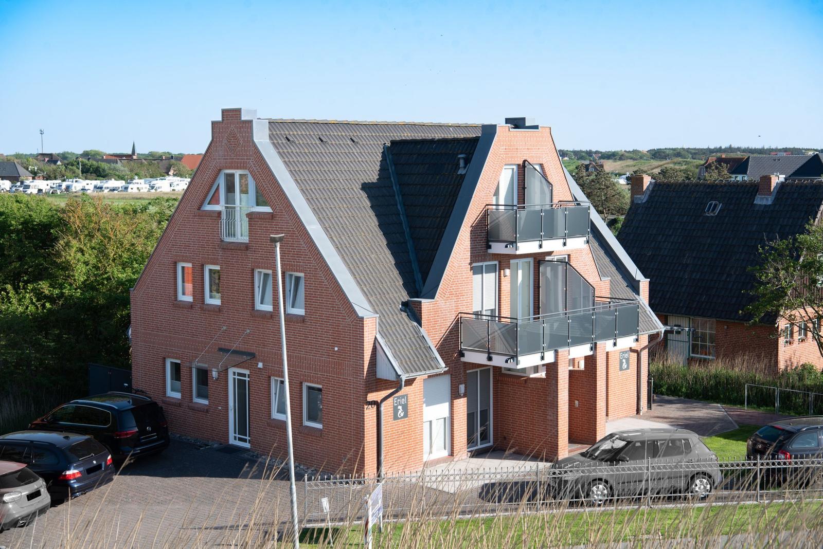 Zweistöckiges Haus mit Balkonen und Parkplatz. Im Hintergrund sind Wohnmobile und Bäume zu sehen.