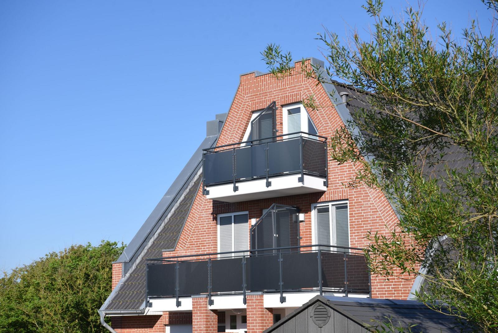 Zwei Balkone mit schwarzen Geländern an einem roten Ziegelhaus unter blauem Himmel.