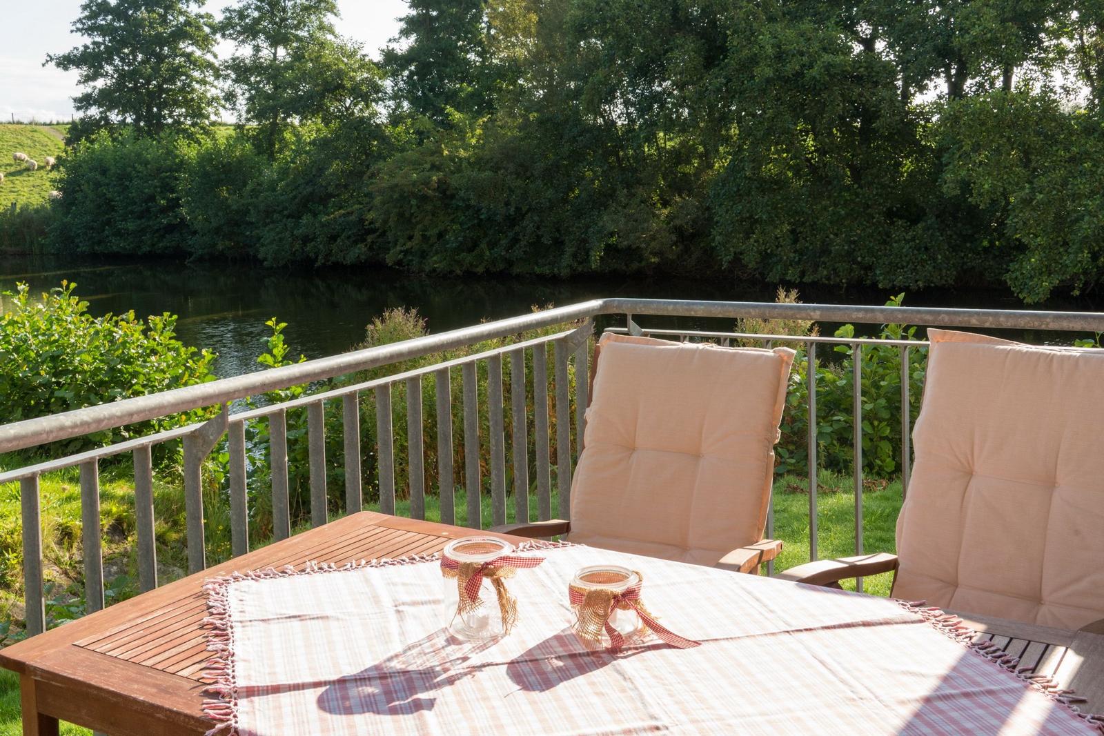 Terrasse mit Tisch und zwei Stühlen, Blick auf einen Fluss und grüne Bäume.
