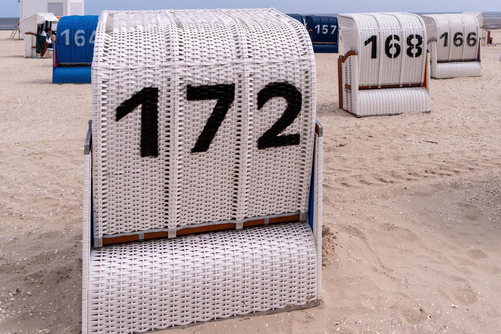 Weißer Strandkorb mit der Nummer 172 auf dem Sand. Im Hintergrund sind weitere Strandkörbe zu sehen.