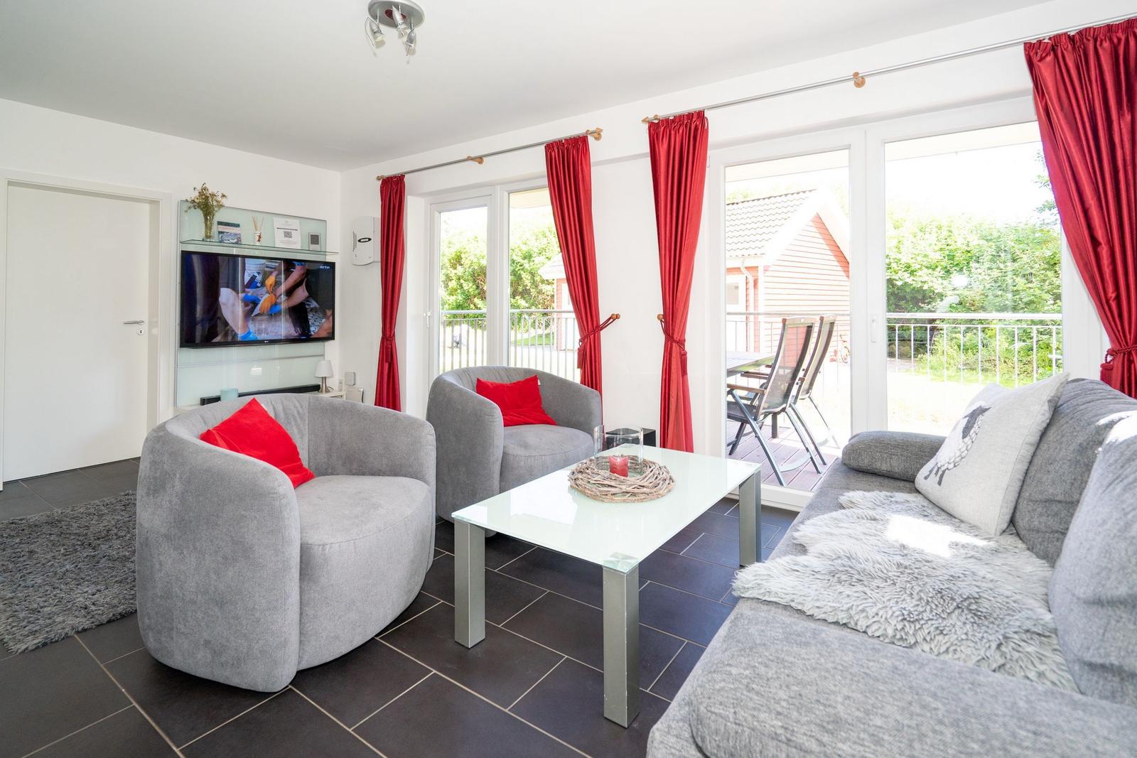 Wohnzimmer mit grauer Couch, roten Vorhängen und Glas-Tisch. Fernseher an Wand. Terrasse mit Blick.