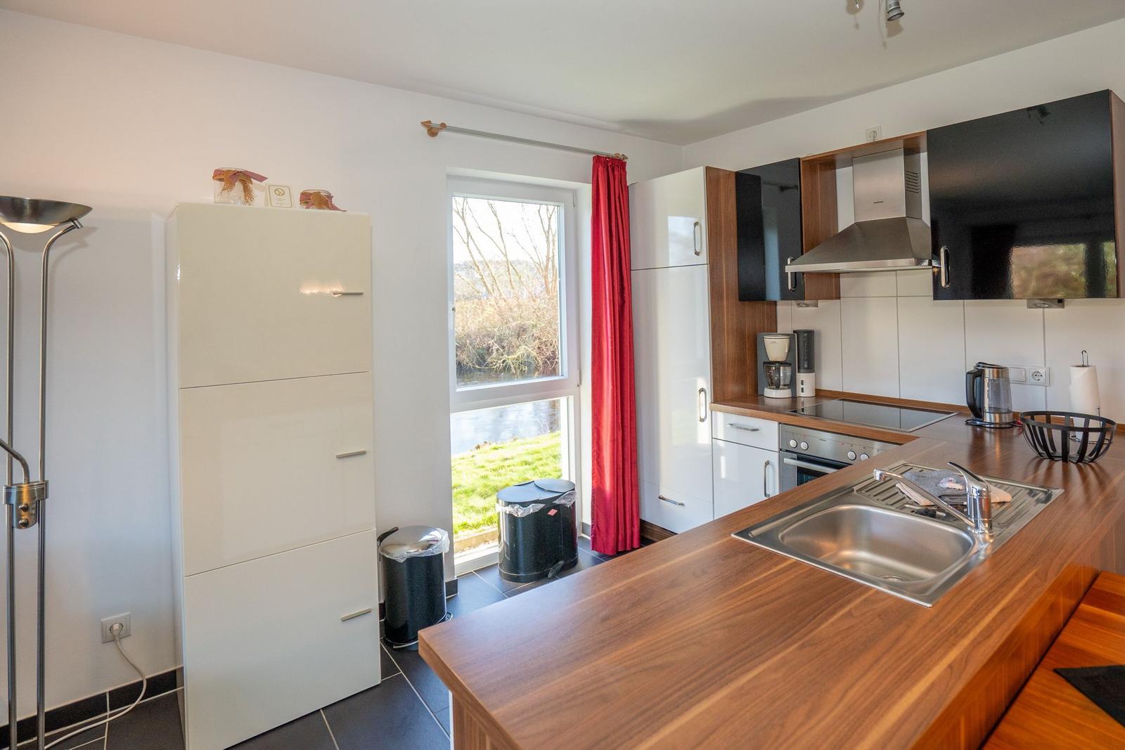 Küche mit Holzoberfläche, Fenster mit Blick auf Wasser, rote Vorhänge und weiße Schränke.