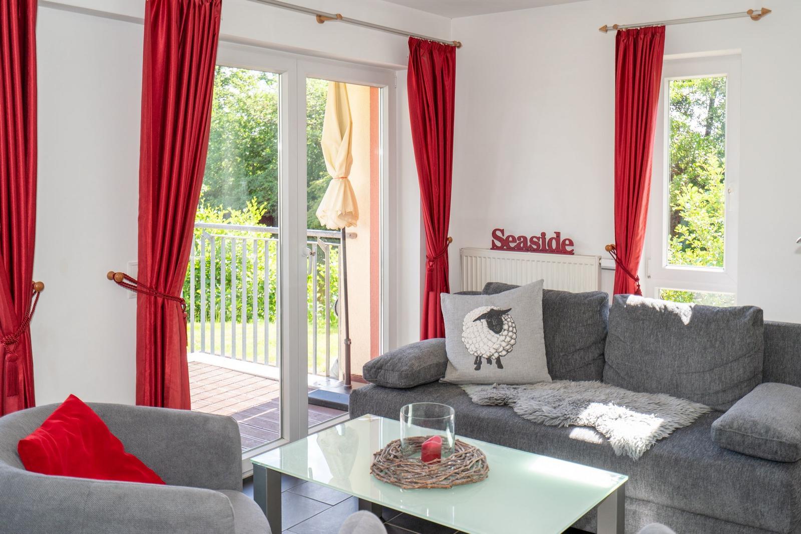 Wohnzimmer mit grauem Sofa, roten Vorhängen und Blick auf Balkon.