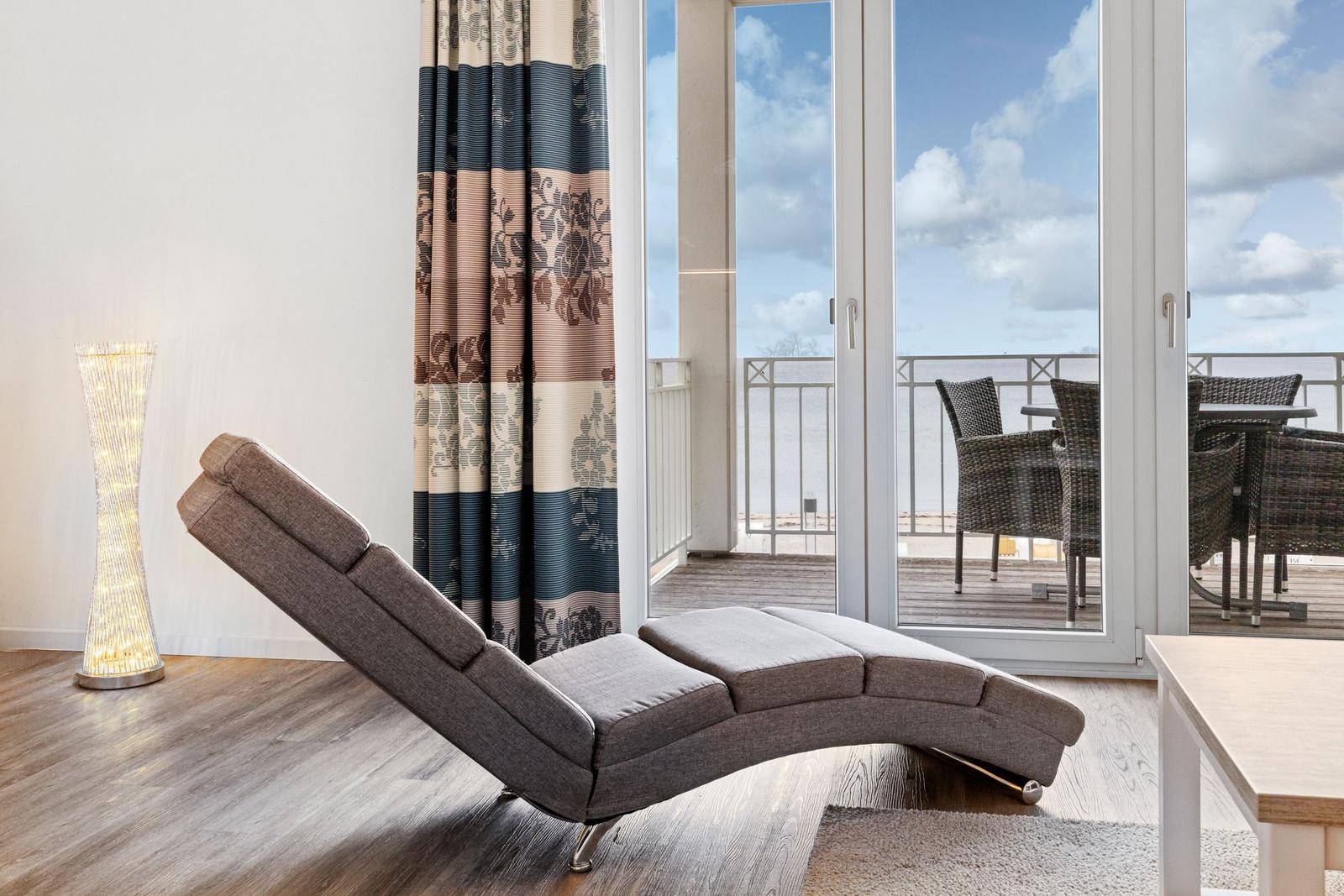 Ein grauer Liegesofa steht vor einer Glastür zum Balkon mit Meerblick.