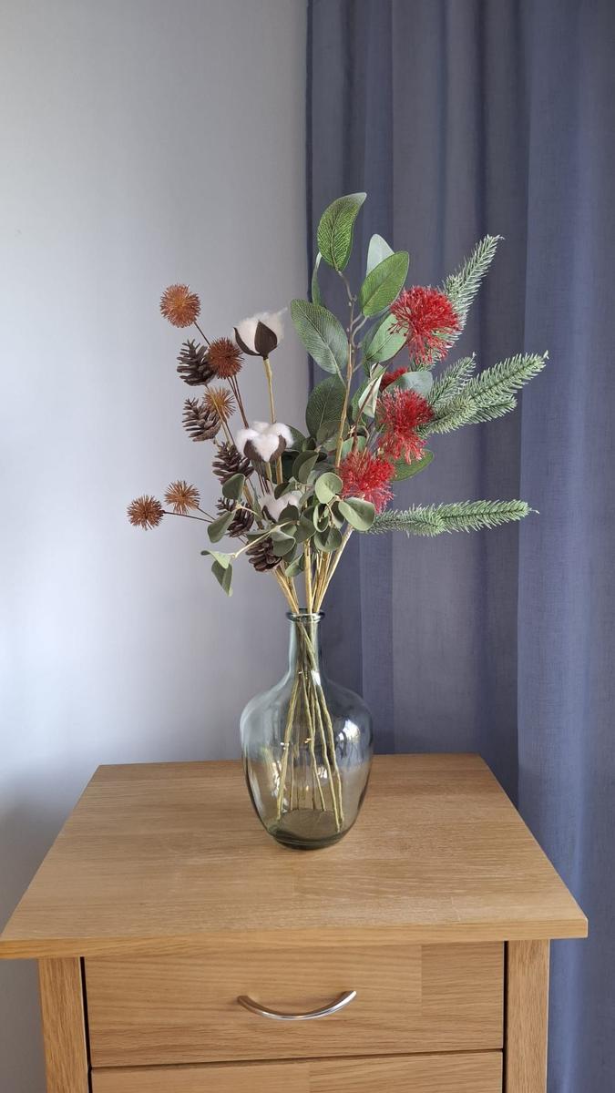 On a wooden dresser, a glass vase holds dried flowers and green leaves.