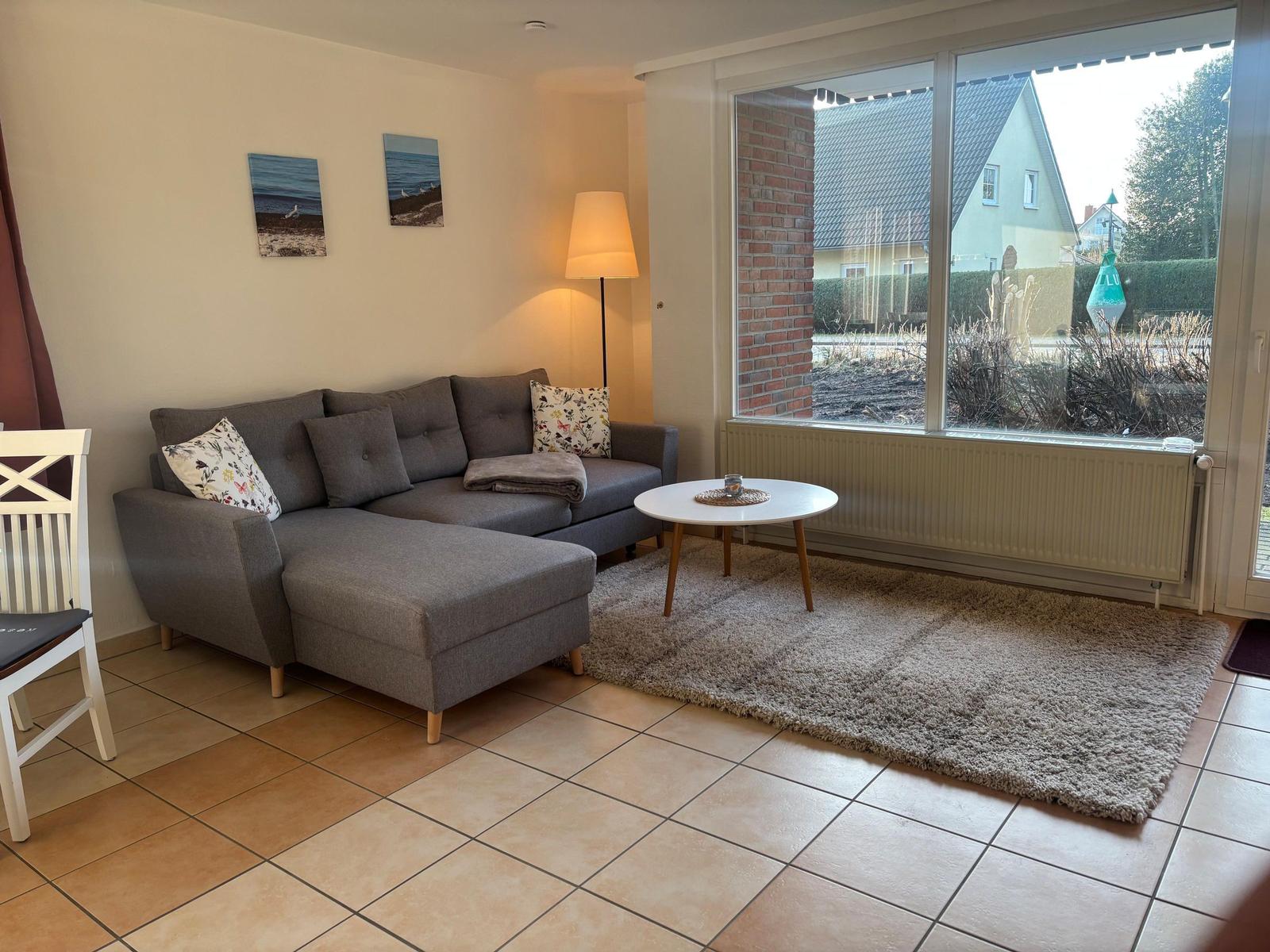 Ein Wohnzimmer mit grauem Sofa, runden Tisch und großem Fenster zur Außenseite.