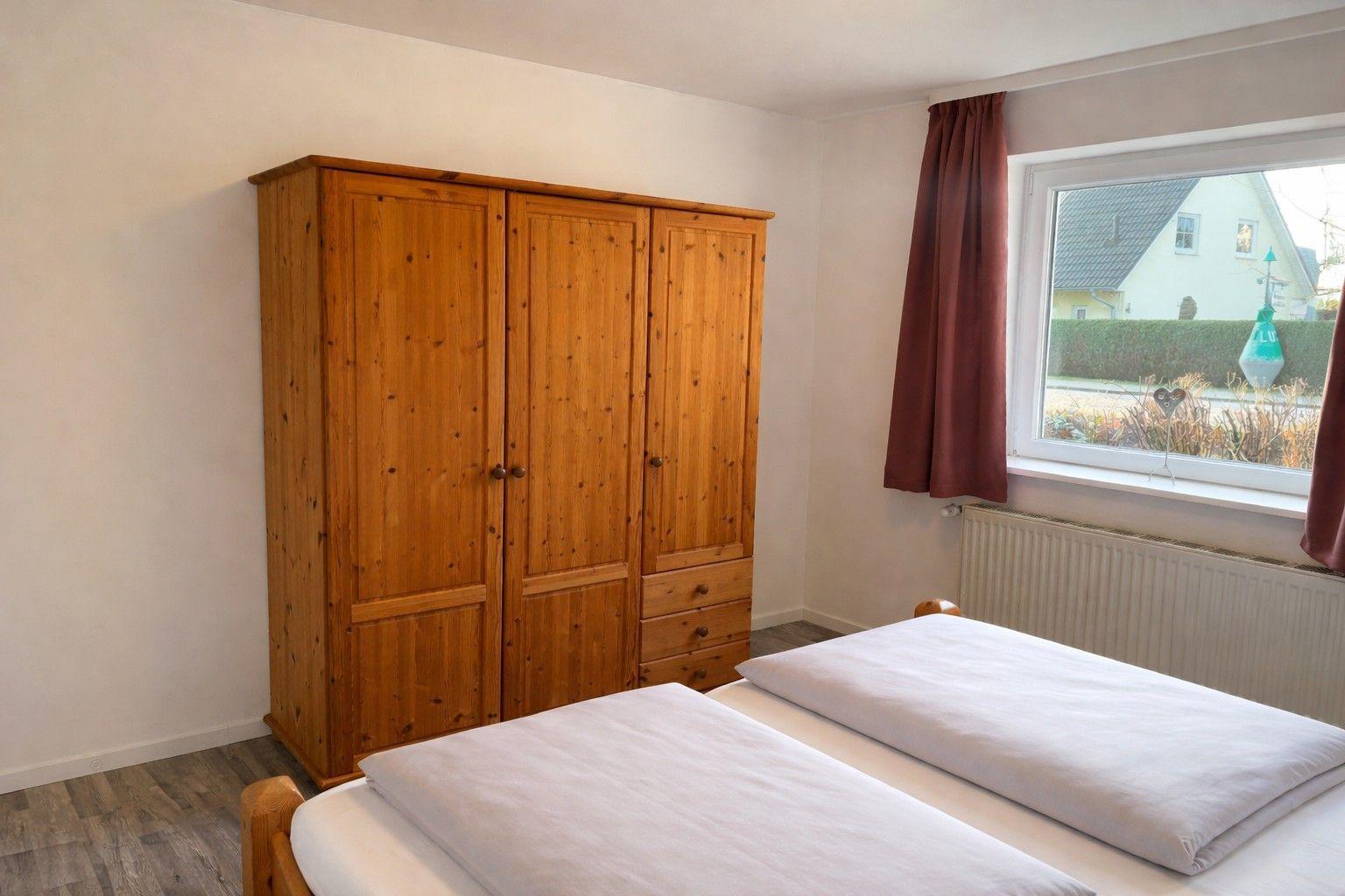 Schlafzimmer mit Holzschrank und Doppelbett. Fenster mit Vorhang. Blick auf Haus und Straße.