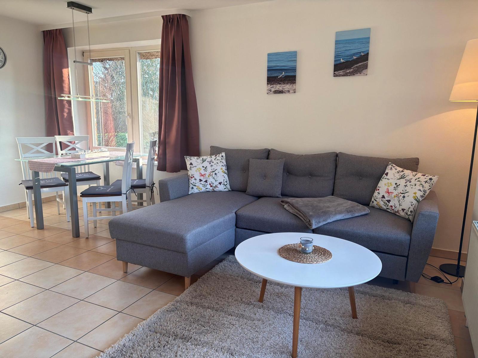 Wohn- und Essbereich mit grauem Sofa, runden Tisch und Fenster mit Vorhängen.