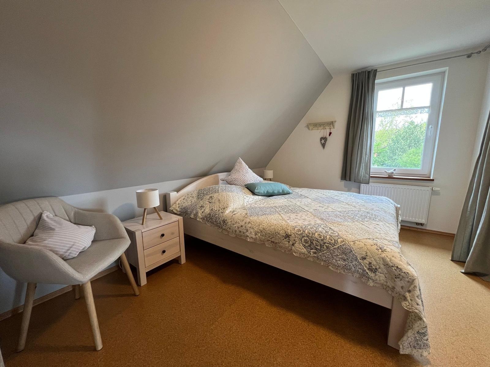 Schlafzimmer mit Dachschräge, Bett, Stuhl und Fenster mit Blick auf Grün.
