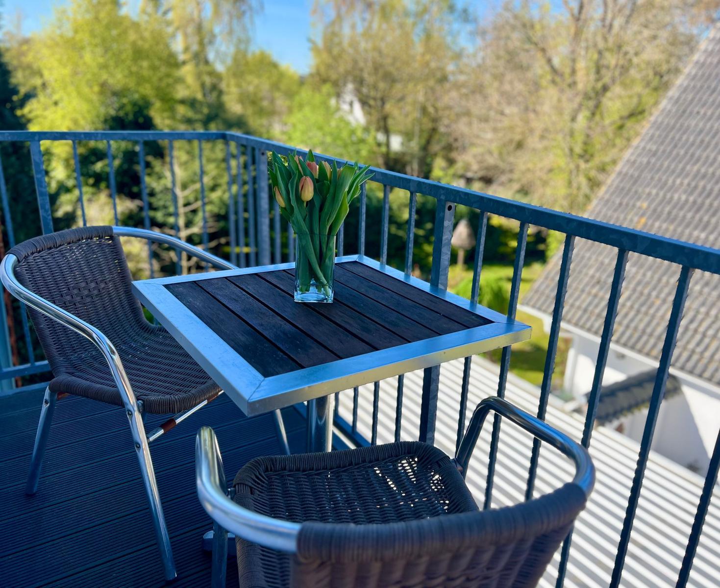 Terrasse mit Tisch und zwei Stühlen, darauf Blumen in Glas. Blick auf grüne Bäume und Dach.