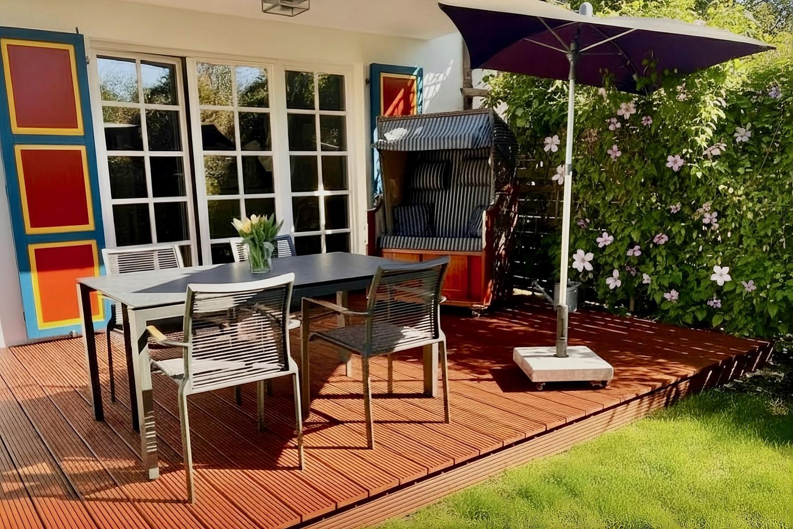 Terrasse mit Tisch, Stühlen, Sonnenschirm und Liegestuhl vor Haus mit Fenstern.