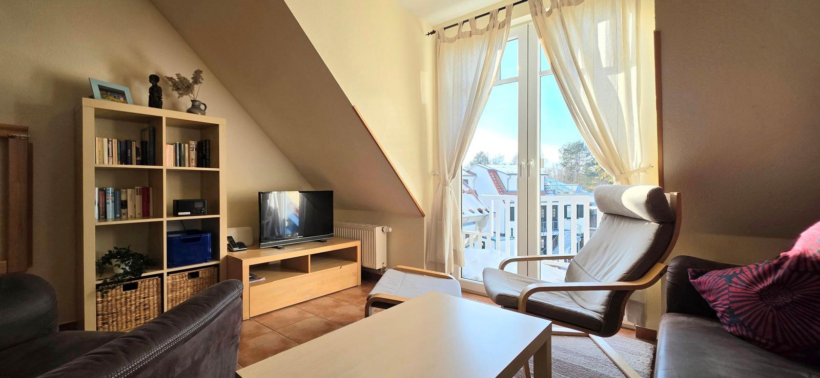 Wohnzimmer mit Fernseher, Bücherregal und Sitzgelegenheiten. Fenster mit Blick auf Balkon.