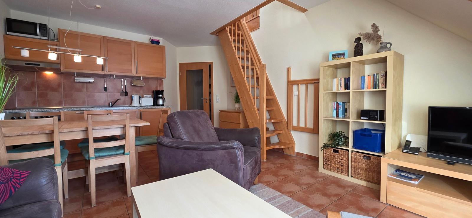 Wohnung mit Küche, Essbereich, Sofa und Bücherregal. Holztreppe führt nach oben.