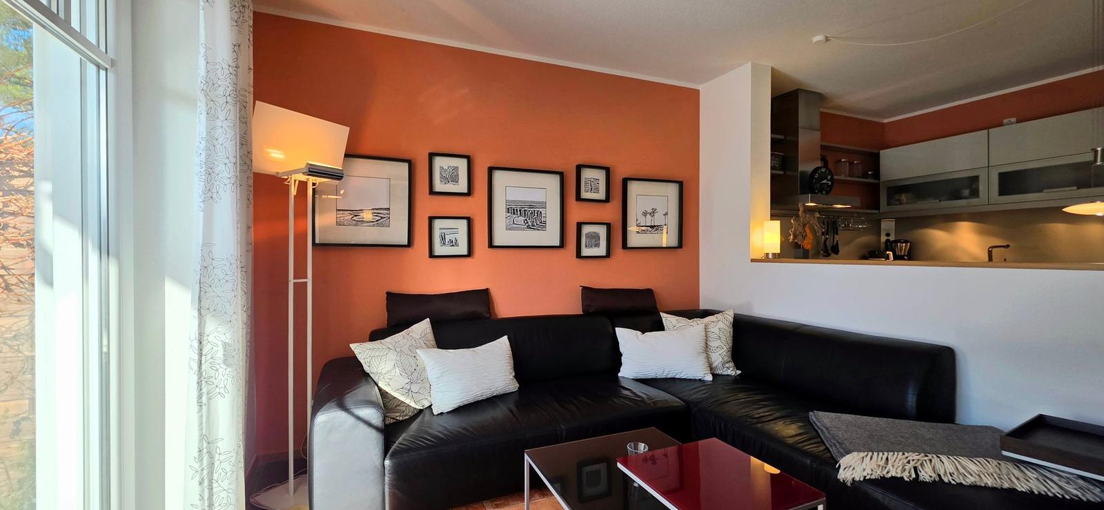 Wohnbereich mit schwarzer Couch, orangefarbener Wand und Küchenbereich im Hintergrund.