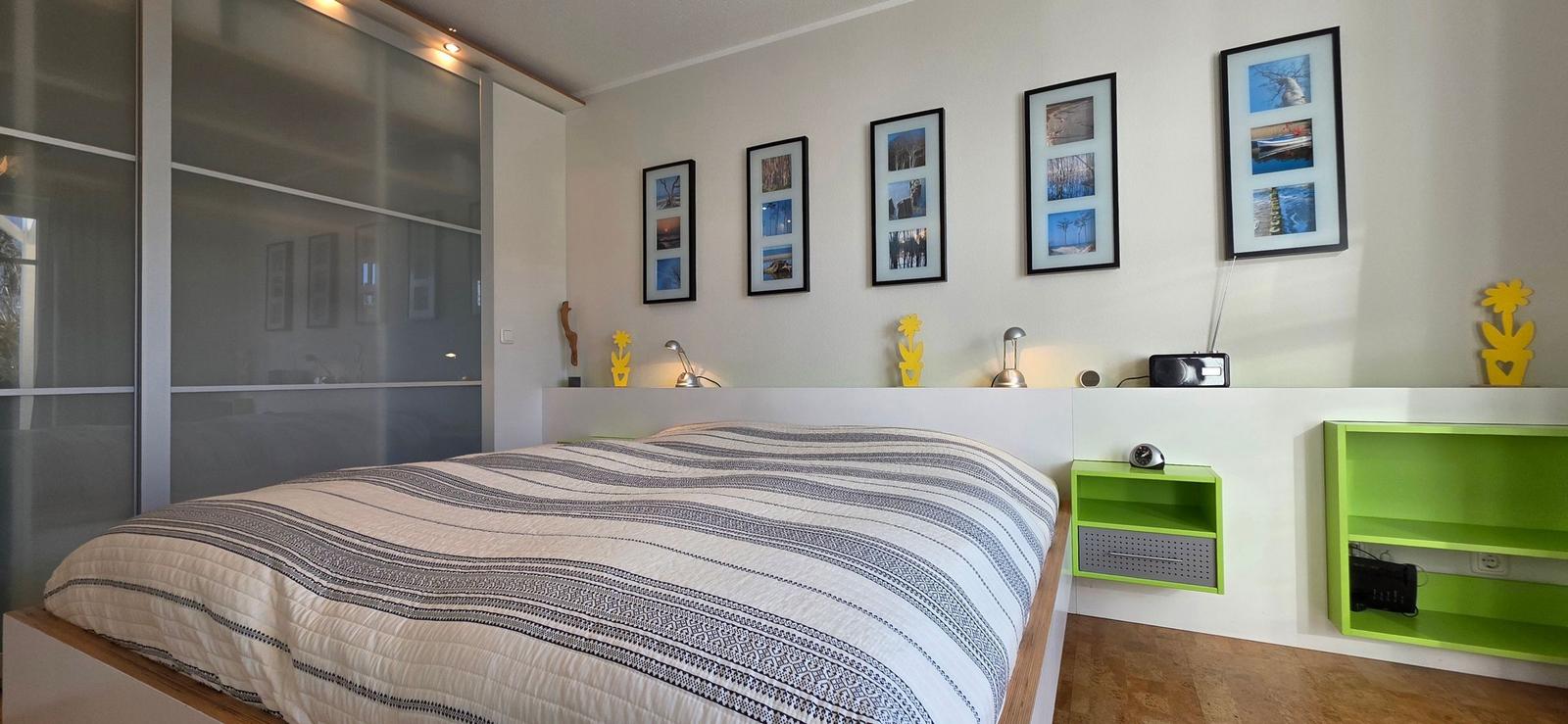 Schlafzimmer mit Bett, Wandbildern und grünen Regalen.