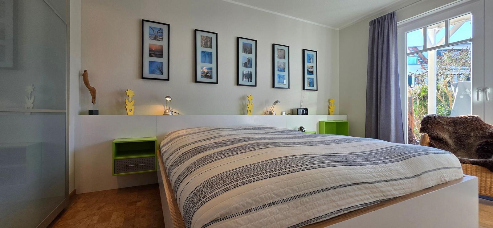 Schlafzimmer mit Bett, Wandbildern und Fenster zur Außenterrasse.