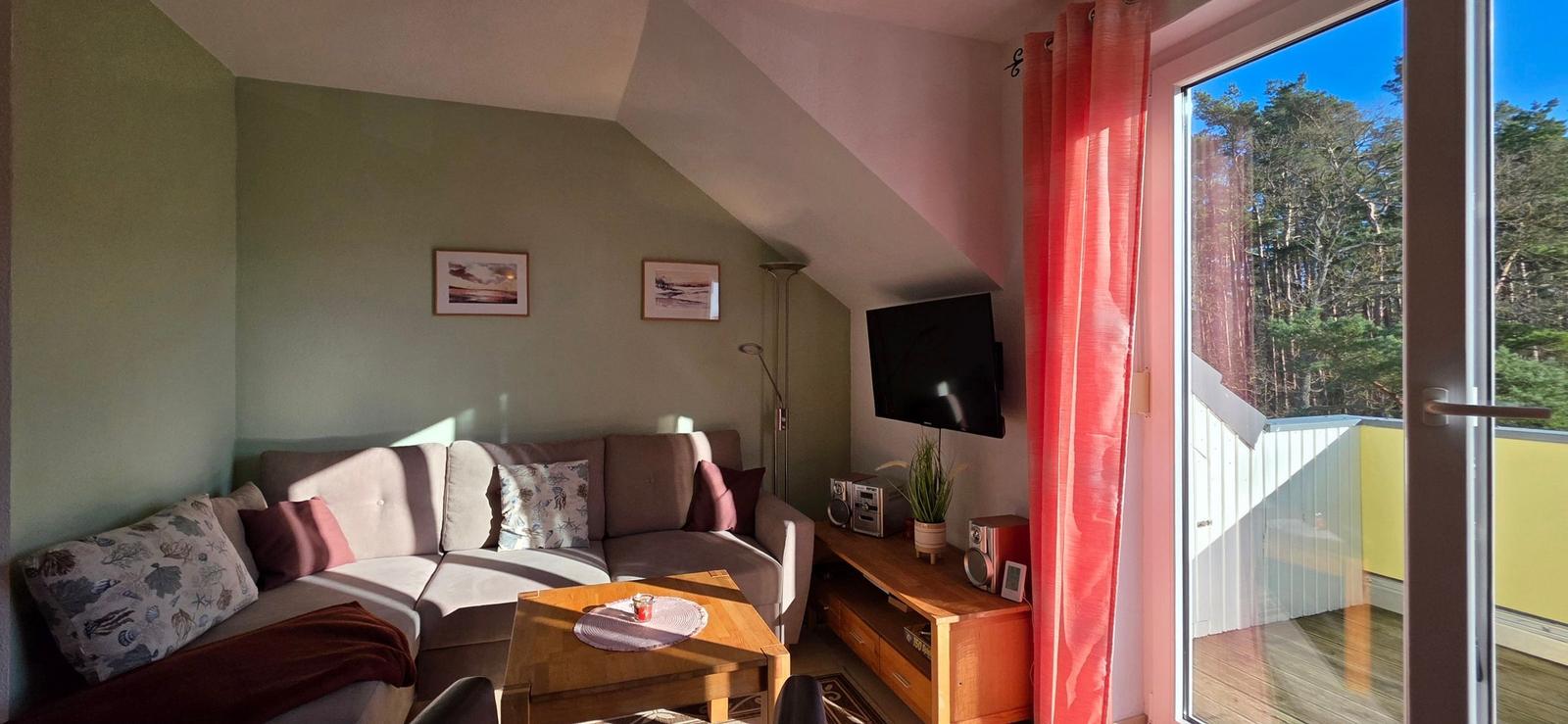 Wohnzimmer mit Sofa, Fernseher und Balkon mit Blick auf Wald.