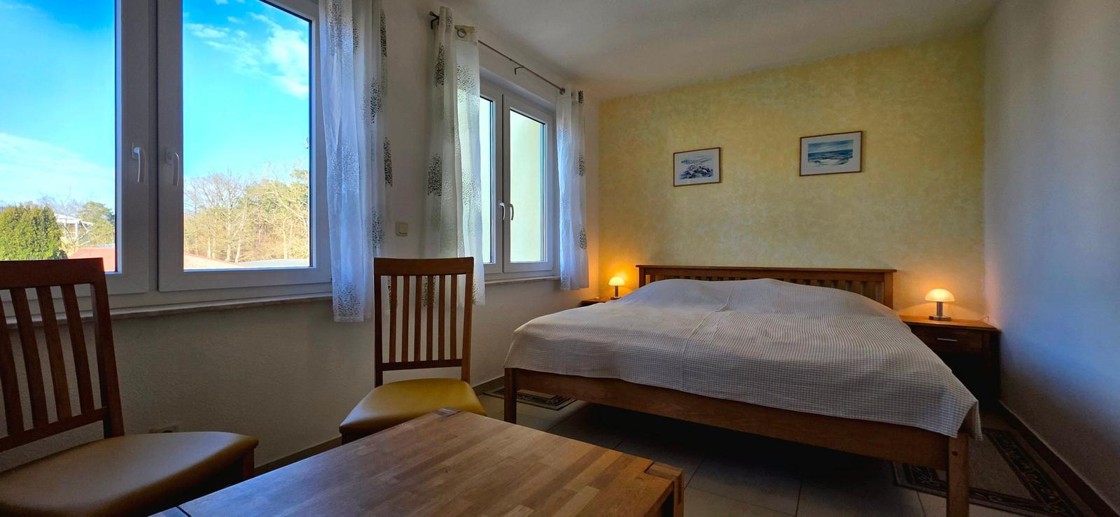 Schlafzimmer mit Bett, Tisch, Stühlen und Fenstern mit Blick auf Außenbereich.