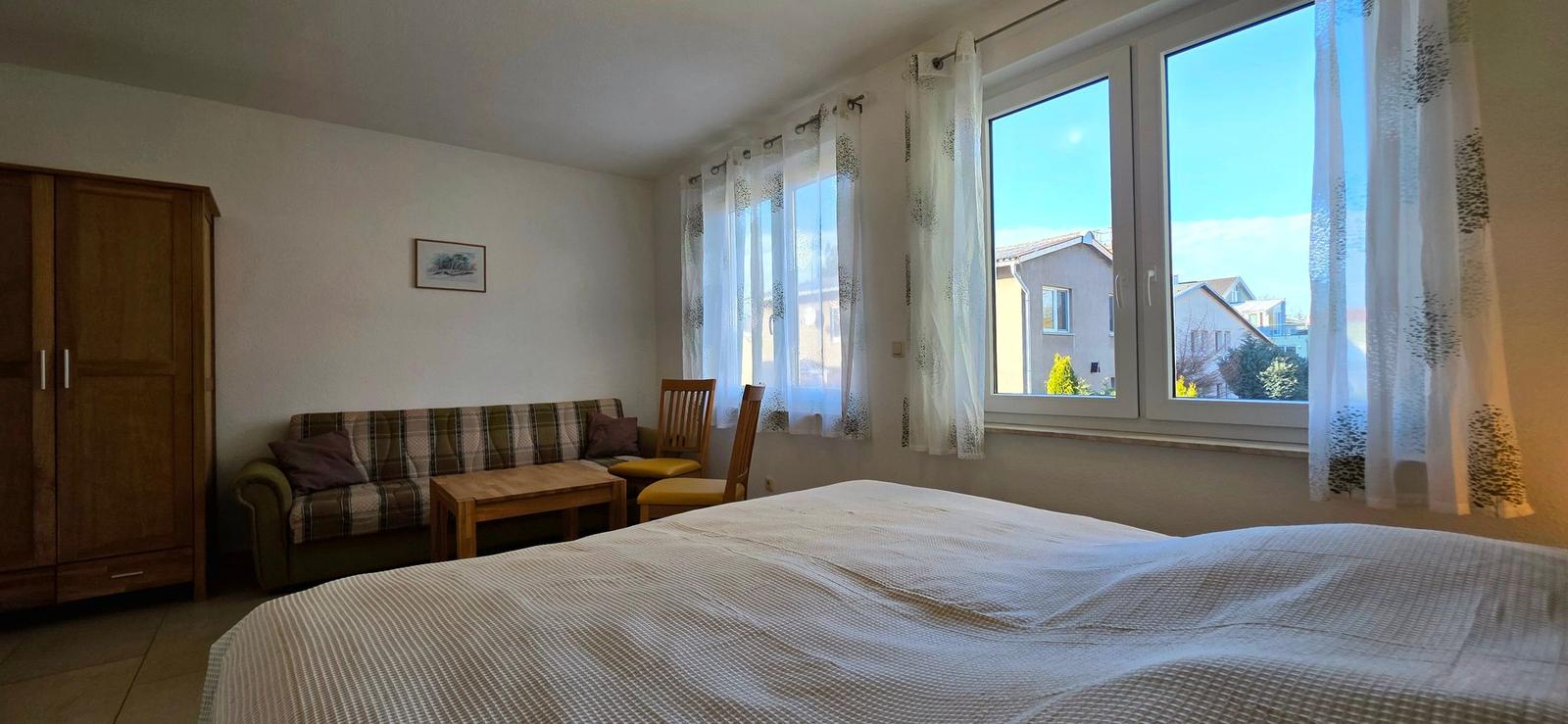 Zimmer mit Bett, Sofa, Tisch und Stühlen. Fenster mit Blick auf Wohngebiet.