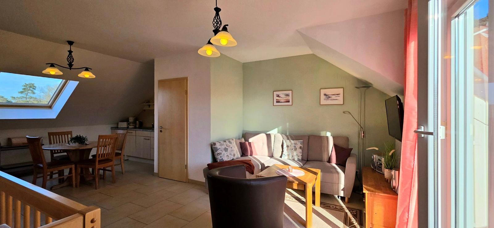 Wohn- und Essbereich mit Sofa, Tisch und Fenster. Holzmöbel und Deckenleuchten.