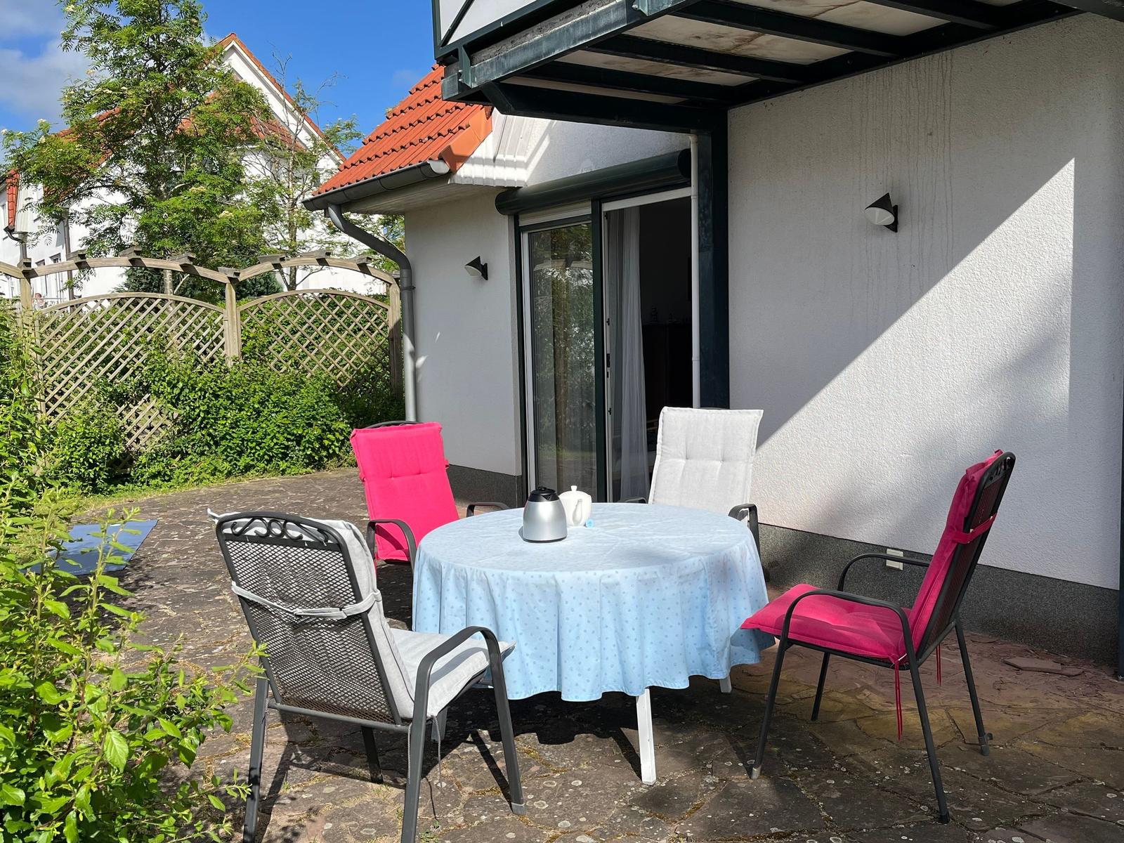 Terrasse mit runden Tisch und vier Stühlen unter Pavillon. Hintergrund: Garten mit Zaun und Haus.