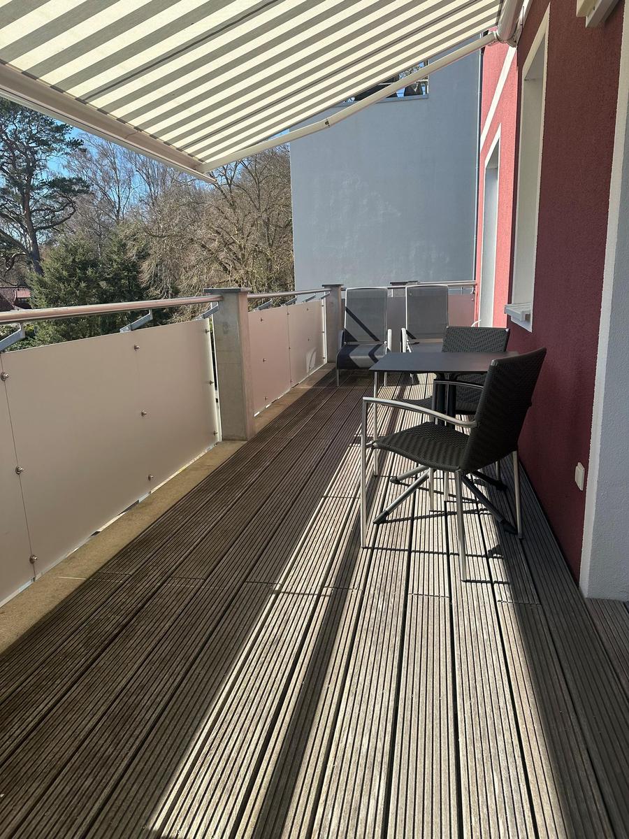 Terrasse mit Tisch, Stühlen und Markise. Holzdielen und Sicht auf Bäume.