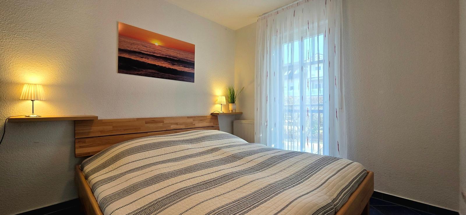 Schlafzimmer mit Bett, Wandbild und Fenster mit Vorhängen.