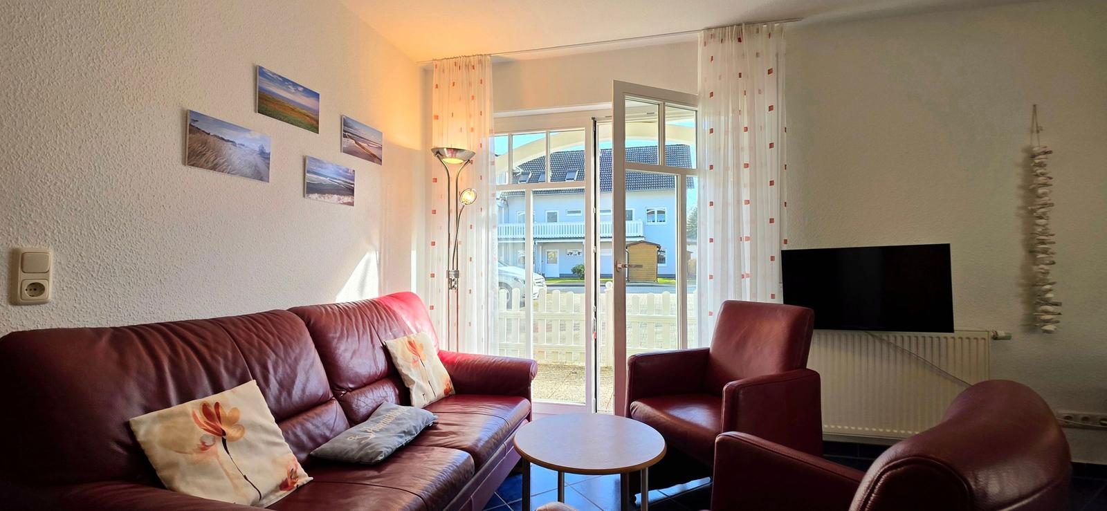 Wohnzimmer mit rotem Sofa, zwei Sesseln, Fernseher und Blick auf den Hof.