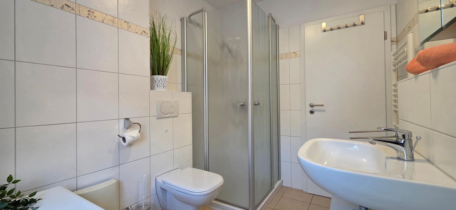 Bathroom with shower, sink, and toilet. White tiles and plants.