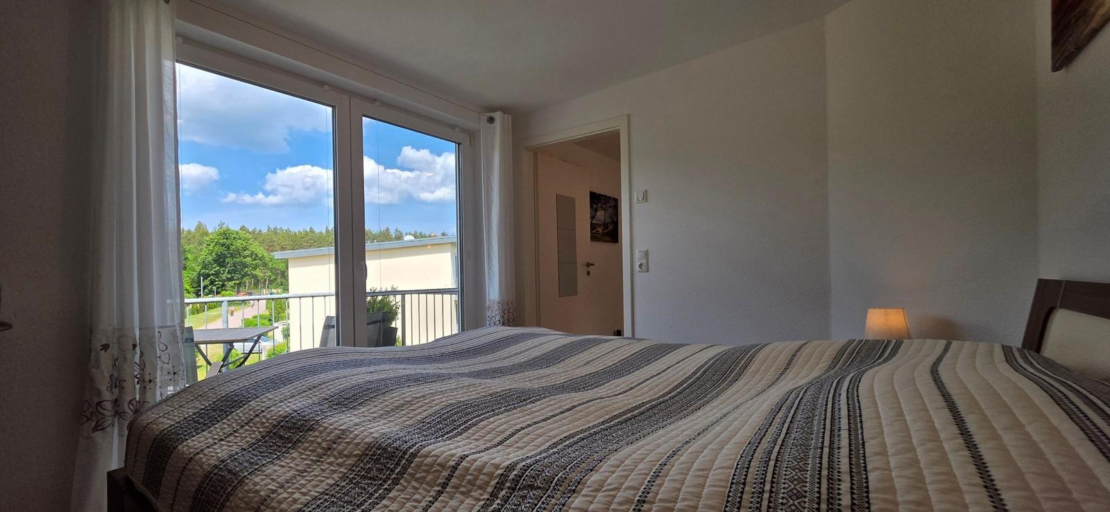 Schlafzimmer mit Blick auf Balkon und Wald. Bett mit Streifenbezug.