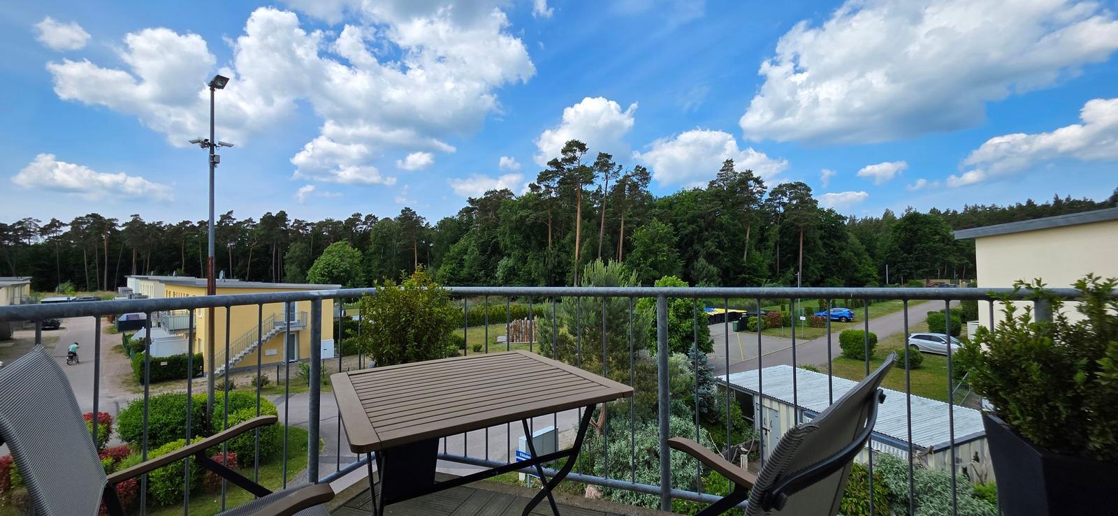 Terrasse mit Tisch und Stühlen, Blick auf Wald und Wohnanlage unter blauem Himmel.