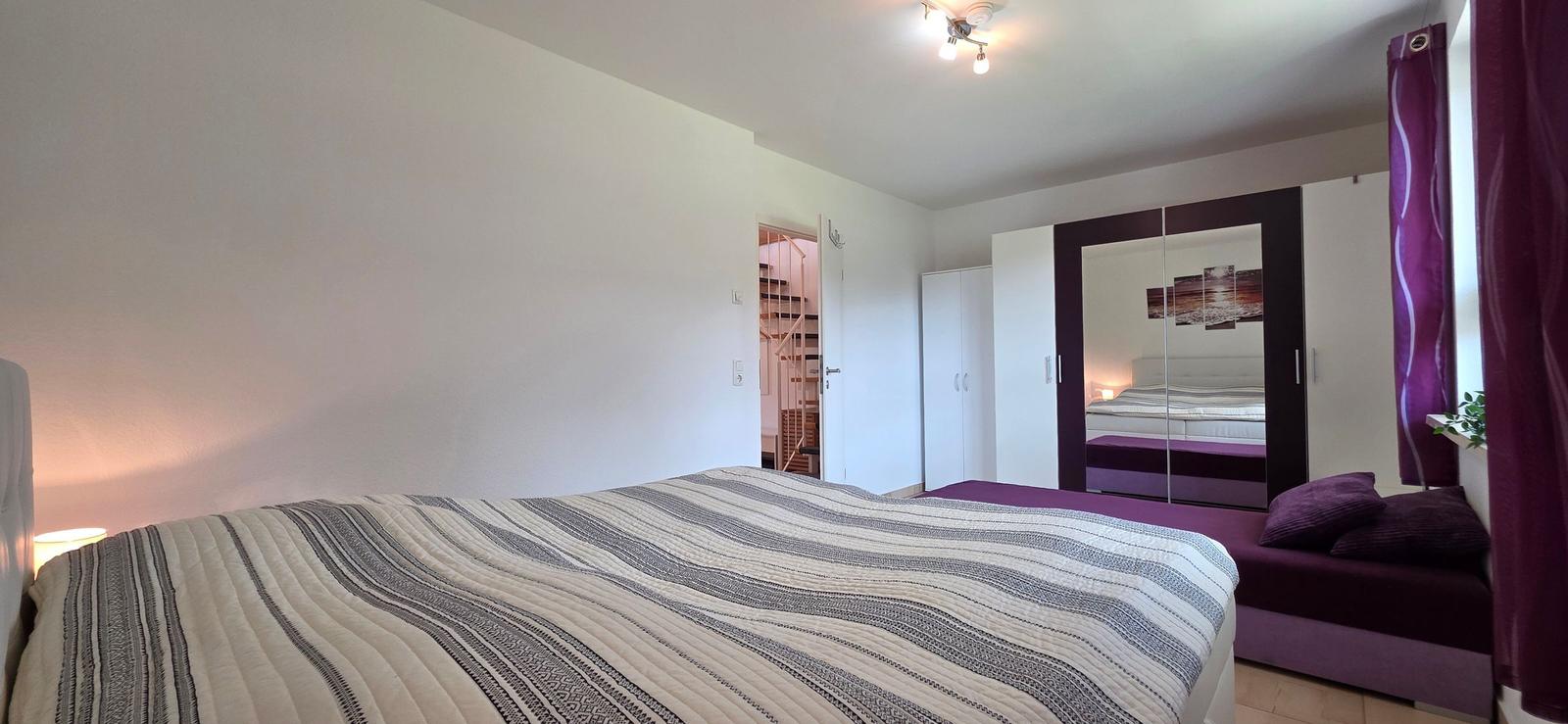 Schlafzimmer mit Bett, Kleiderschrank und Sitzbank. Durchgang zur Treppe sichtbar.