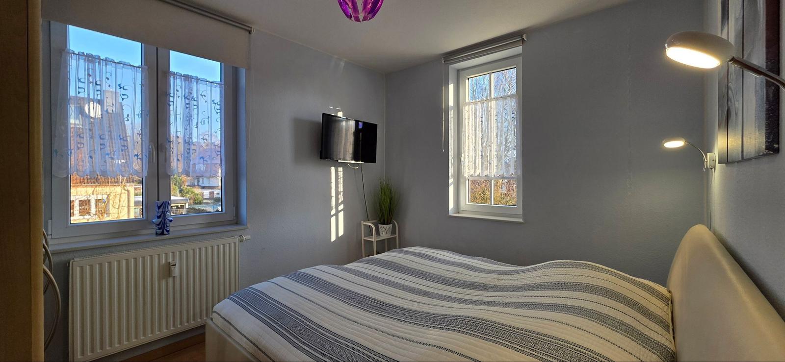 Schlafzimmer mit Bett, TV und zwei Fenstern. An der Wand hängt eine Lampe.